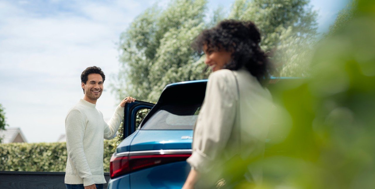 Man opening blue car door. Smiling to woman walking towards him. 