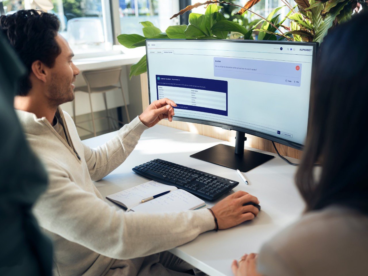 Person sitting at a desk in a modern office, interacting with data on a widescreen monitor, with a notebook and keyboard on the table and green plants in the background.